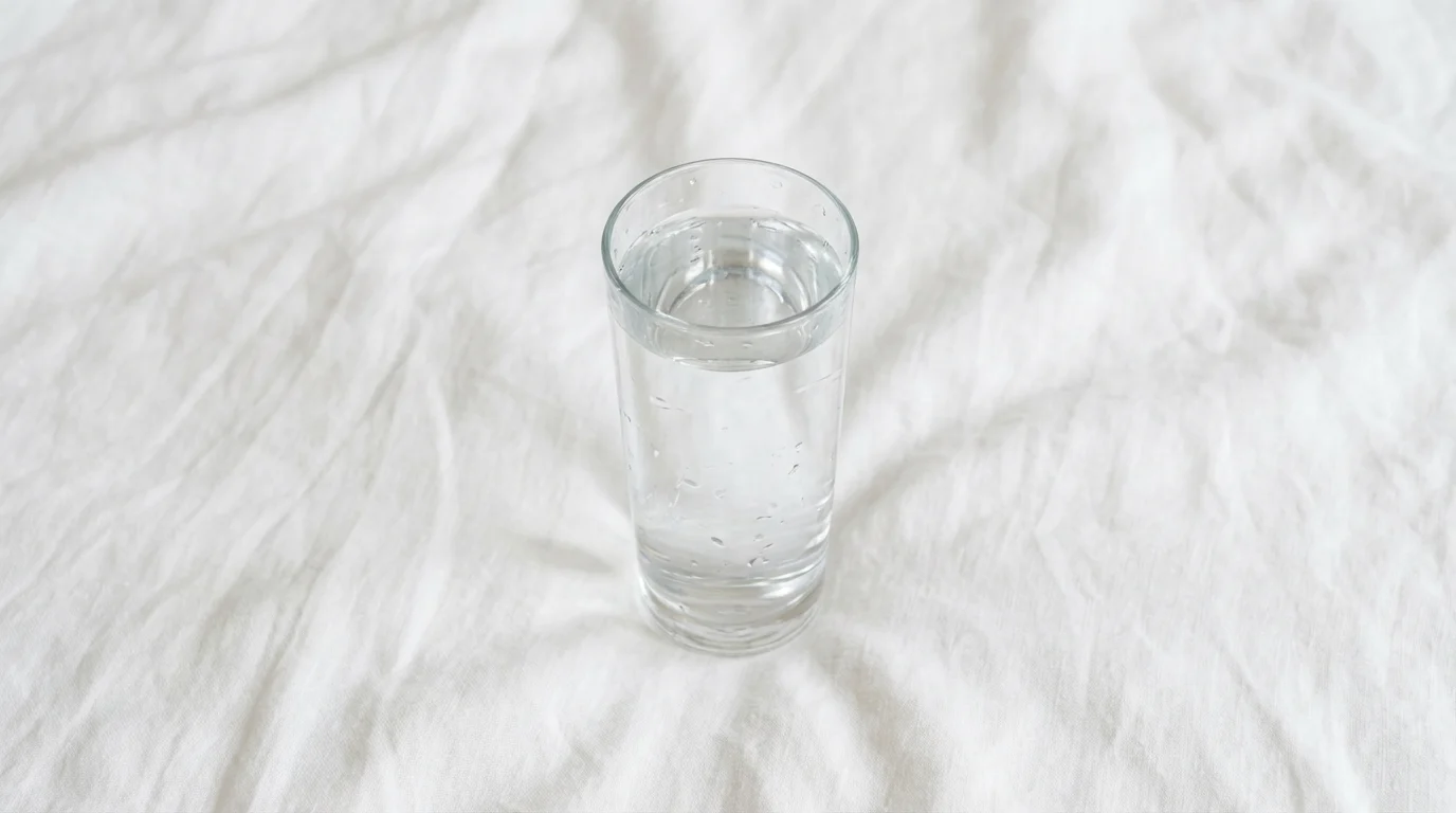High angle flat lay photograph of a condensation-covered glass of water on white bed linen.