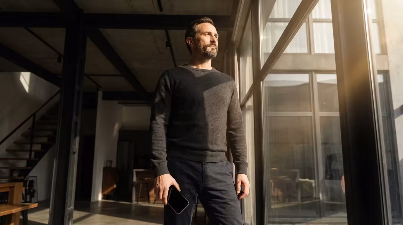 Man standing by window holding phone at side loosely with black screen