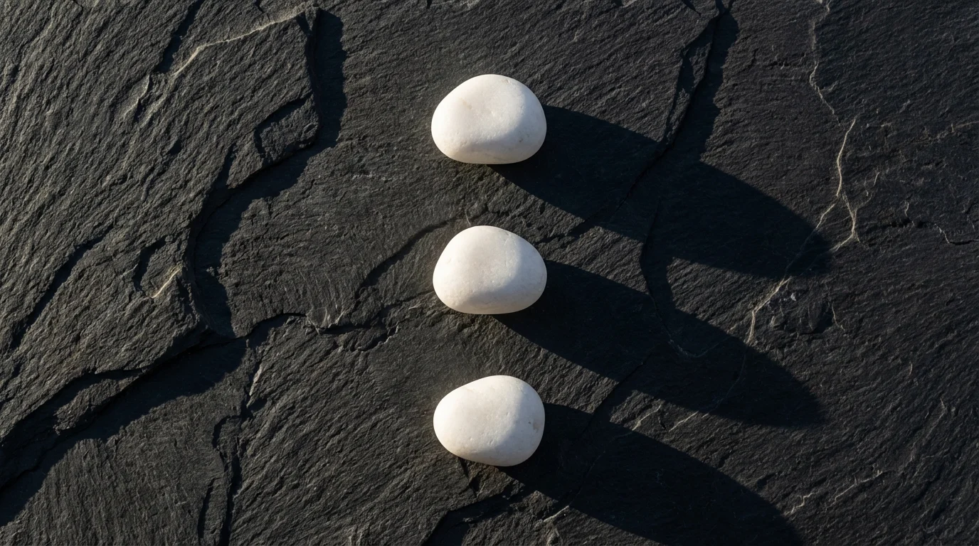 Overhead view of three white stones lined up on dark slate with dramatic afternoon shadows.